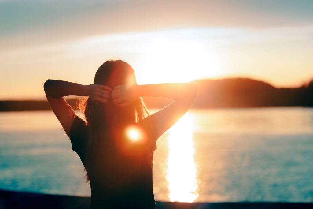 Woman with hands behind head watching the sunset over the water